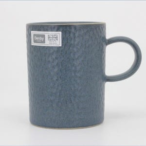 May include: A blue ceramic mug with a textured, dimpled surface and a single handle. A rectangular white sticker is attached to the upper left side, featuring the text Denby, a small Union Jack flag, and the words Made in England.