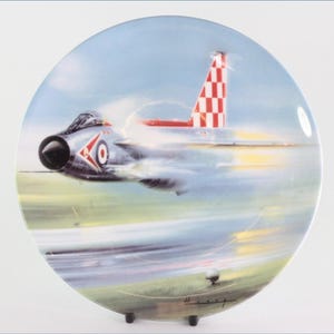 Könnte beinhalten: Ein dekorativer Teller mit einem Düsenflugzeug im Flug. Das Flugzeug ist silberfarben mit rot-weiß karierten Heckflossen. Der Teller hat einen blauen Himmel und einen grünen Landschaftshintergrund.