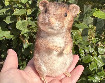 May include: A needle-felted mouse figurine crafted from wool fibers in shades of brown, tan, and cream. The mouse features small rounded ears, dark bead eyes, a pointed snout, and thin whiskers, standing in an upright posture.