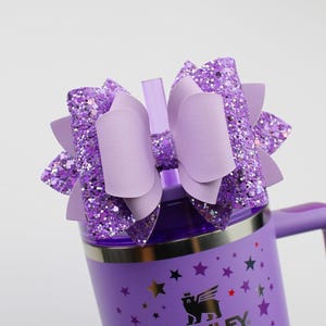 May include: A purple insulated tumbler with a handle, decorated with stars and a logo. It is topped with a large, layered bow in shades of purple and glitter. A clear straw is inserted into the lid.