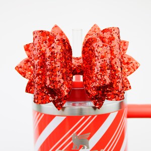 May include: A red glitter bow is attached to a red and white striped tumbler. The bow is made of layered fabric and is a vibrant shade of red with sparkling glitter. The tumbler has a red handle.