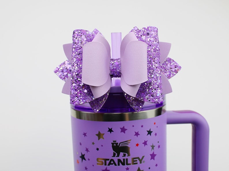 May include: A purple tumbler with a matching bow and straw. The bow is made of glitter and solid purple material. The tumbler has a handle and a logo with the text "STANLEY". The tumbler is decorated with stars.