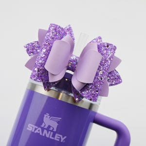 May include: A purple Stanley tumbler with a matching handle and a decorative bow. The bow is made of solid and glittery lavender material. A clear straw is inserted into the tumbler's lid.