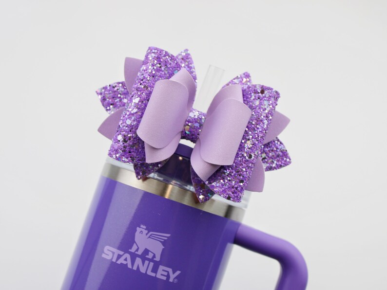 May include: A purple Stanley tumbler with a matching handle and a decorative bow. The bow is made of solid and glittery lavender material. A clear straw is inserted into the tumbler's lid.