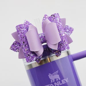 May include: A purple Stanley tumbler with a matching bow. The bow is made of glittery purple material and has a solid purple center. A clear straw is inserted into the tumbler's lid. The tumbler has a handle.