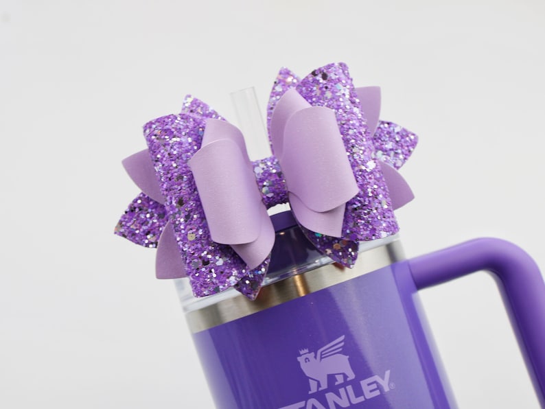May include: A purple Stanley tumbler with a matching bow. The bow is made of glittery purple material and has a solid purple center. A clear straw is inserted into the tumbler's lid. The tumbler has a handle.