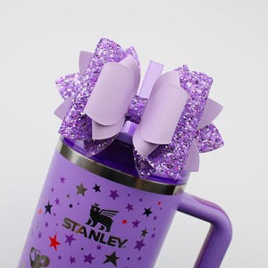 May include: A purple Stanley tumbler with a matching handle and a decorative bow. The bow is made of glittery purple material and solid purple accents. The tumbler has a silver rim and a black logo with stars.