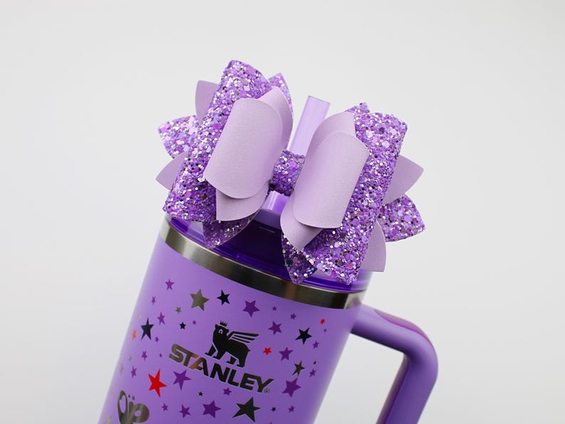 May include: A purple Stanley tumbler with a matching handle and a decorative bow. The bow is made of glittery purple material and solid purple accents. The tumbler has a silver rim and a black logo with stars.