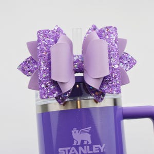 May include: A purple Stanley tumbler with a matching bow. The bow is made of glitter and solid purple material. The tumbler has a handle and a straw. The Stanley logo is visible on the front of the tumbler.