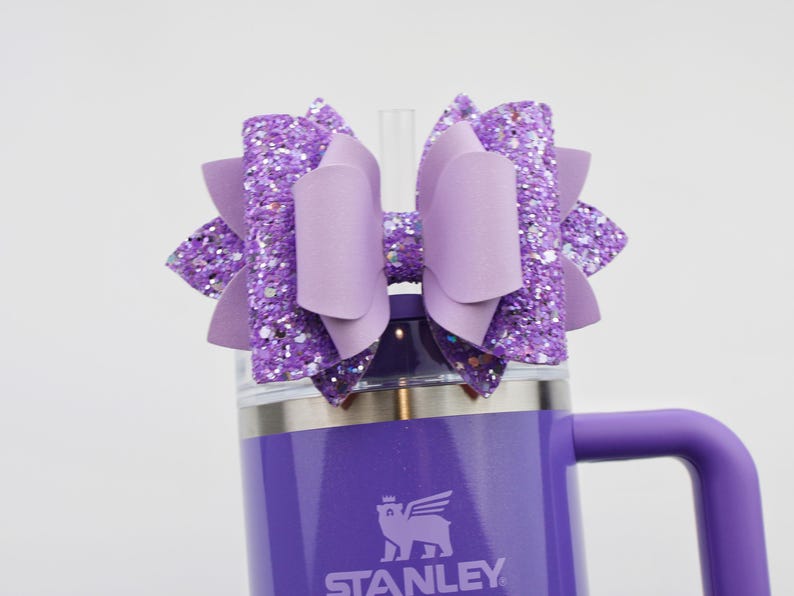 May include: A purple Stanley tumbler with a matching bow. The bow is made of glitter and solid purple material. The tumbler has a handle and a straw. The Stanley logo is visible on the front of the tumbler.