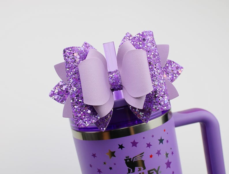 May include: A purple insulated tumbler with a handle, decorated with stars and a logo. It is topped with a large, layered bow in shades of purple and glitter. A clear straw is inserted into the lid.