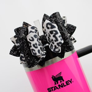 May include: A bright pink Stanley tumbler with a black handle, topped with a decorative bow. The bow features black glitter and leopard print fabric. A clear straw is inserted into the tumbler.