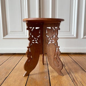 Op de afbeelding: Een kleine, ronde houten plantenstandaard met ingewikkelde gesneden bloemen- en bladerontwerpen op de poten. De standaard is gemaakt van warm getint hout en heeft een driepotige basis. De bovenkant is een solide, cirkelvormig oppervlak.