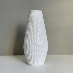 以下が含まれることがあります： テクスチャ加工された幾何学模様の白いセラミック花瓶。花瓶は、上部に向かって狭くなる広いベースを持っています。花瓶の高さは約30cmで、ニュートラルな背景に置かれています。