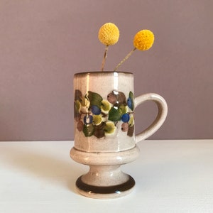 Puede incluir: Una taza de cerámica con base marrón y blanca y un diseño floral con hojas verdes y bayas azules. La taza tiene un asa y una base de pedestal. Dos flores secas amarillas están en la taza.