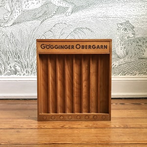 Könnte beinhalten: Eine Holzaufbewahrungsbox mit vertikalen Trennwänden und einer Reihe kleiner kreisförmiger Aussparungen am Boden. Auf der Box steht der Text "GÖGGINGER OBERGARN". Die Box ist aus hellbraunem Holz.