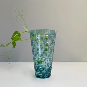 May include: A tall, teal-colored glass vase with a geometric pattern. A green plant with delicate leaves and tendrils extends from the vase. The vase is on a white surface against a gray background. The vase is approximately 8 inches tall.