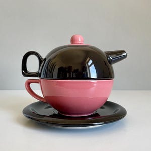 Puede incluir: Un juego de té de cerámica negro y rosa. El juego incluye una tetera con un pomo rosa, una taza de té y un platillo. La tetera se asienta sobre la taza de té, y la taza de té descansa sobre el platillo. El juego tiene un diseño moderno.