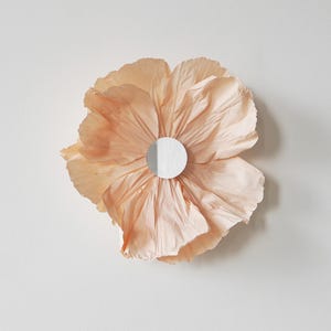 Könnte beinhalten: Eine große, pfirsichfarbene Papierblumen-Wanddekoration mit einem weißen, kreisförmigen Mittelteil. Die Blume hat mehrere gerüschte Blütenblätter und ist an einer weißen Wand befestigt.