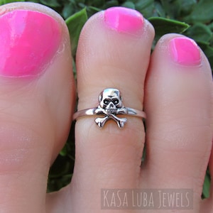 May include: A silver toe ring with a skull and crossbones design. The ring is worn on a person's toe with pink nail polish.