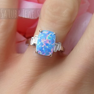 May include: A silver ring with a large blue opal gemstone and two smaller clear gemstones on the band. The ring is on a finger.