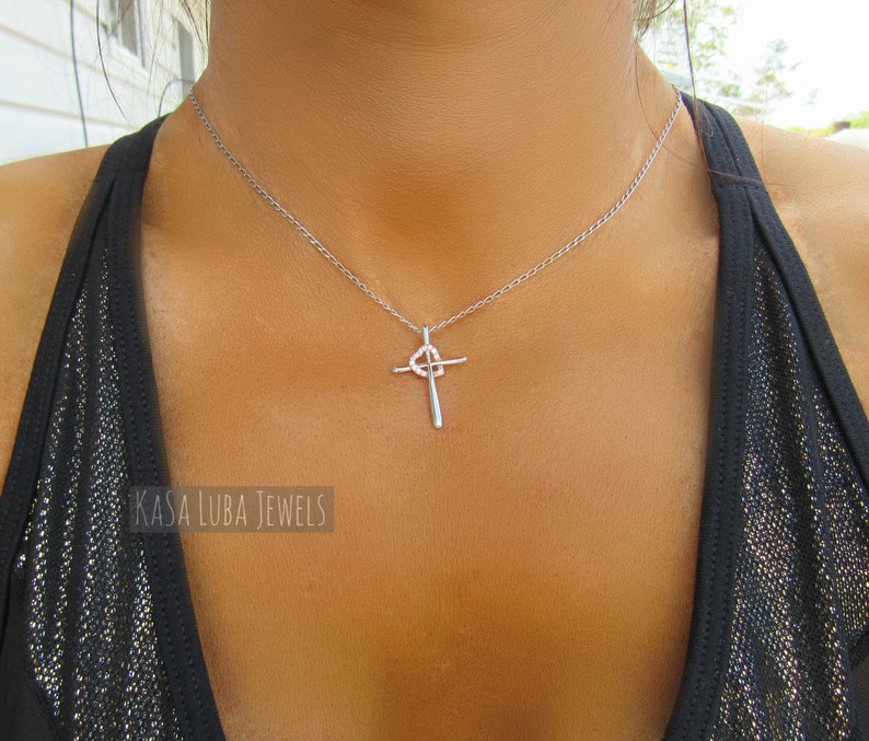 Puede incluir: Un collar de cadena de plata con un colgante de cruz de plata. La cruz tiene un peque&ntilde;o c&iacute;rculo en el centro.