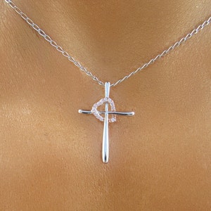 Puede incluir: Un collar de cadena de plata con un colgante de cruz de plata. La cruz tiene una forma de coraz&oacute;n en el centro con peque&ntilde;as piedras transparentes.