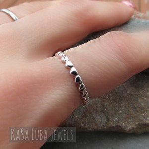 May include: A silver ring with a repeating heart design. The ring is worn on a finger, with the Kasa Luba Jewels logo visible in the lower left corner. The ring is made of a shiny metal and is in focus.