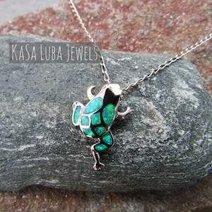 May include: A silver frog pendant necklace with a green and black opal design. The frog is detailed with small legs and a chain. The text "KASA LUBA JEWELS" is visible in the image.