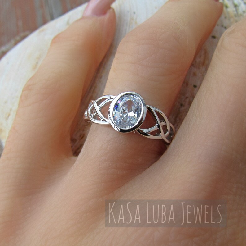 K&ouml;nnte beinhalten: Ein silberner Ring mit einem gro&szlig;en, ovalen, klaren Edelstein. Das Band hat ein kompliziertes keltisches Knotenmuster. Der Ring wird an einer Hand gezeigt. Der Text "KASA LUBA JEWELS" befindet sich unten.