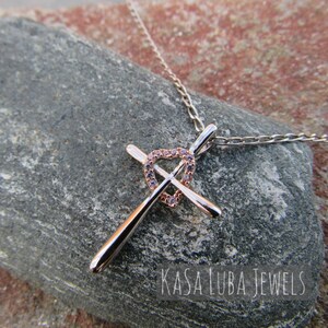 Puede incluir: Un collar de plata y oro rosa con un colgante de cruz en forma de coraz&oacute;n incrustado con piedras de circonita c&uacute;bica transparentes. El colgante est&aacute; en una cadena de plata. El collar est&aacute; sobre una roca gris. El texto "Kasa Luba Jewels" es visible en la imagen.