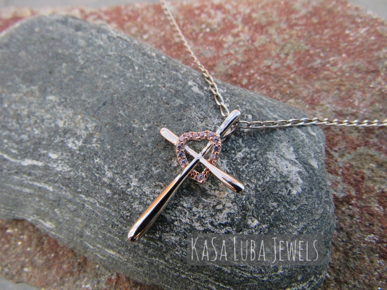 Puede incluir: Un collar de plata y oro rosa con un colgante de cruz en forma de coraz&oacute;n incrustado con piedras de circonita c&uacute;bica transparentes. El colgante est&aacute; en una cadena de plata. El collar est&aacute; sobre una roca gris. El texto "Kasa Luba Jewels" es visible en la imagen.