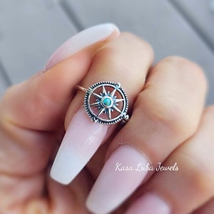 May include: A silver compass ring with a turquoise stone in the center. The ring is on a finger with long, white acrylic nails. The text "Kasa Luba Jewels" is visible in the bottom right corner of the image.