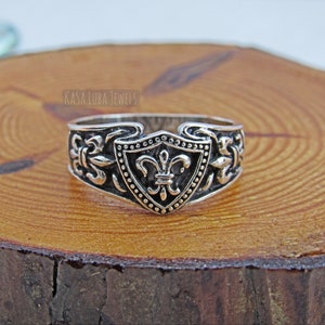 May include: A silver ring with an ornate design. The ring features a shield-shaped centerpiece with a fleur-de-lis symbol. The band is decorated with floral and scrollwork patterns. The ring is displayed on a wooden surface.
