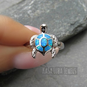May include: A silver ring with a blue opal turtle design. The ring is on a finger and the background is a gray stone.