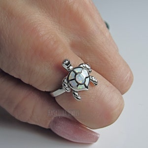May include: A silver-toned ring featuring a turtle design. The turtle's shell is inlaid with a white opal-like material, and the ring is detailed with textured patterns. The ring is on a finger, with the brand name "KASA LUBA JEWELS" visible.