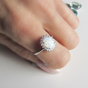 May include: A silver ring with a white opal gemstone surrounded by a halo of smaller clear stones. The ring is on a hand with pink nail polish.