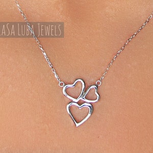 May include: Silver necklace with a three-heart pendant. The hearts are interlocking and have a delicate, open design. The necklace is on a model's neck.