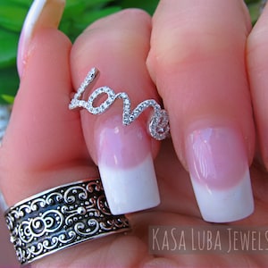 May include: A silver ring with the word "LOVE" spelled out in cubic zirconia stones. The ring is on a finger with a white and pink french manicure.