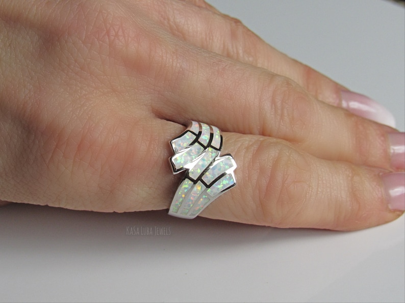 Op de afbeelding: Een zilveren ring met een ontwerp van witte opaal. De ring wordt gedragen aan een vinger.