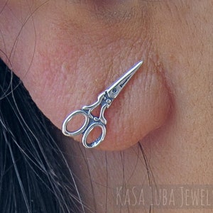 May include: Silver earring featuring a pair of scissors design. The earring is shown in a person's ear.