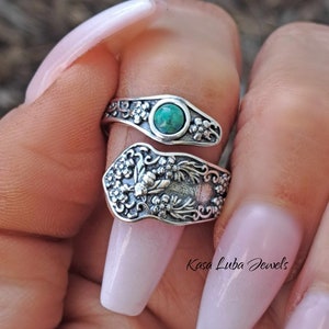 May include: A silver ring with a green gemstone and a bee design. The ring is wrapped around a finger and has a floral pattern.