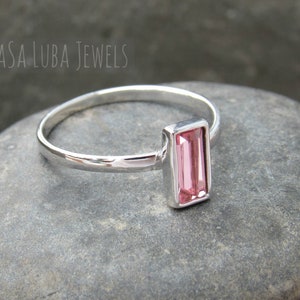 May include: A silver ring with a pink rectangular gemstone. The ring is on a gray stone surface.
