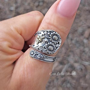 May include: A silver and gold thumb ring with a floral design featuring sunflowers and a bee. The ring is made from a repurposed spoon and is a unique piece of jewelry.
