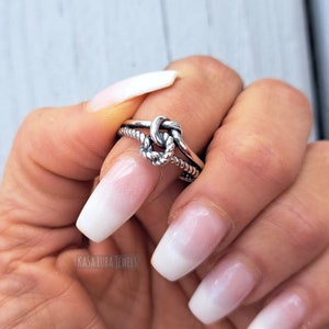 May include: A silver ring with a knot design. The ring is worn on a finger with white nail polish.