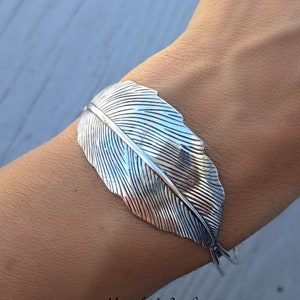 May include: A silver feather cuff bracelet. The feather is textured and detailed. The bracelet is open and adjustable.