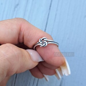May include: A silver knot ring with a simple design. The ring is worn on a finger and the knot is centered on the top of the finger.
