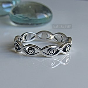 May include: Silver ring with a repeating pattern of stylized eyes. The ring is on a white surface. "Kasa Luba Jewels" is printed in the background.