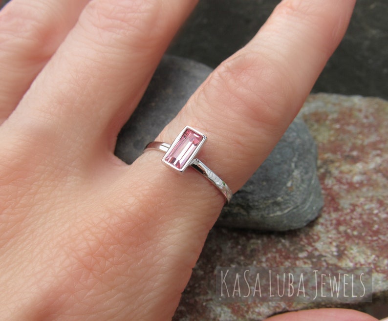 May include: A delicate silver ring with a pink rectangular gemstone set in a bezel setting. The ring band is textured with a hammered finish.