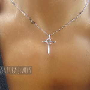 Puede incluir: Un collar de cadena de plata con un colgante de cruz de plata que presenta un peque&ntilde;o dise&ntilde;o de coraz&oacute;n con piedras transparentes. El collar lo lleva una persona con la piel bronceada.
